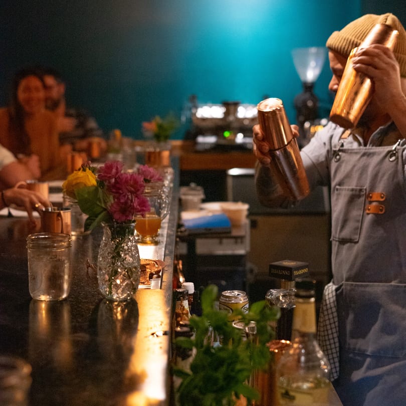 Bartender in a beanie shaking two copper cocktail shakers behind a bar with flowers and drinks.