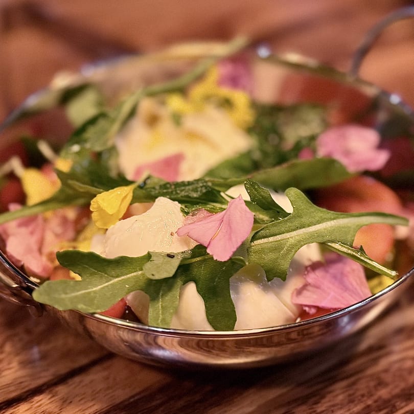 A vibrant salad with burrata, arugula, cherry tomatoes, and edible pink and yellow flowers in a metal bowl.
