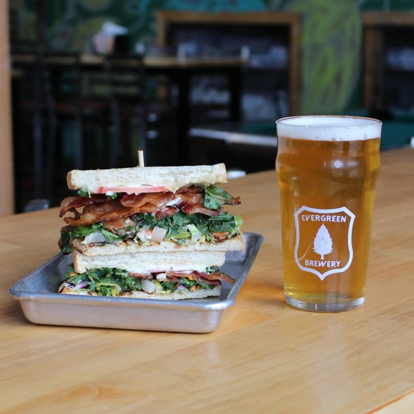 A tall club sandwich on a metal tray next to a pint of Evergreen Brewery beer.