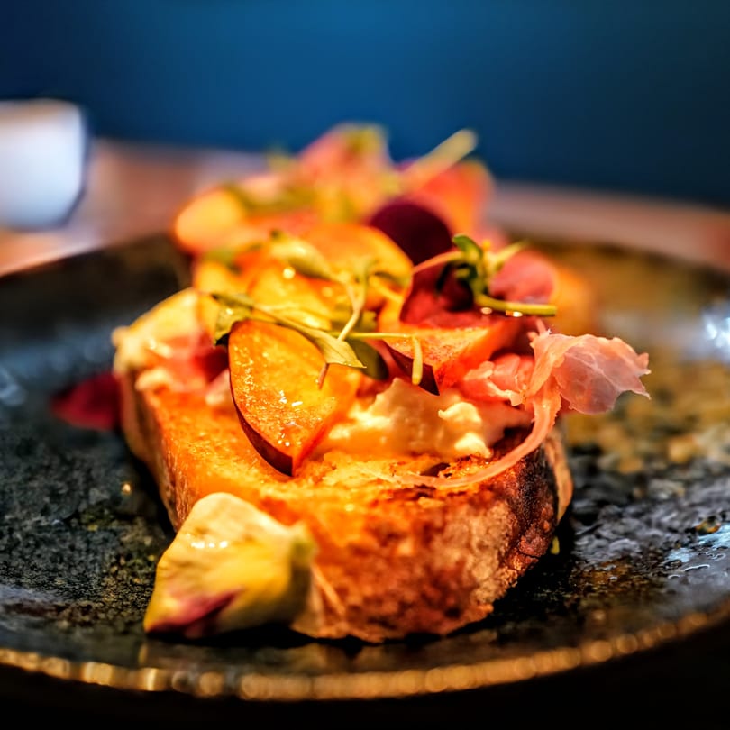 Close-up of bruschetta topped with sliced peaches, prosciutto, and fresh greens on a dark plate.