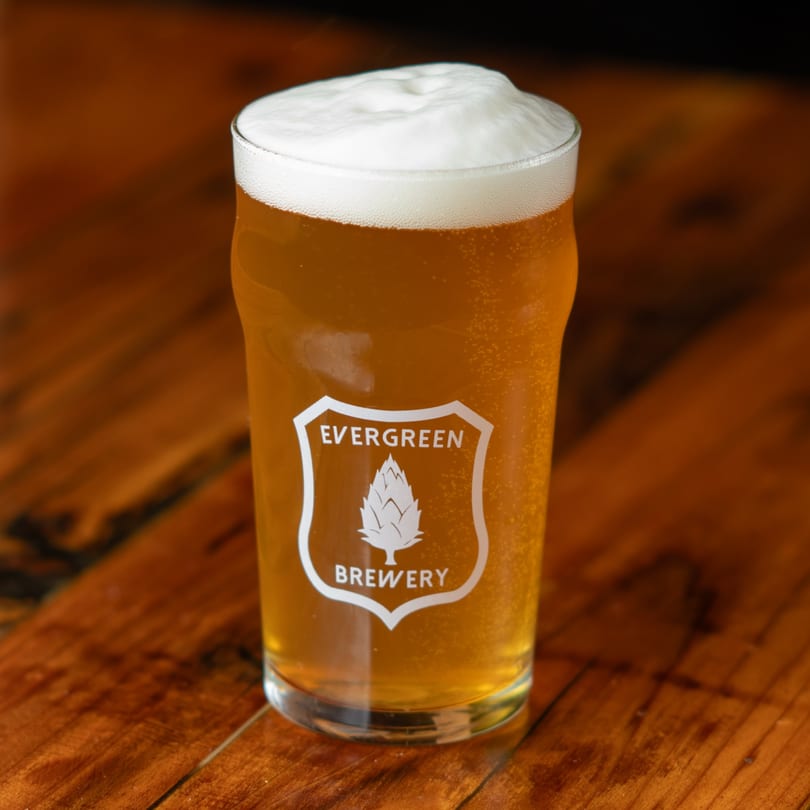 A pint glass of Evergreen Brewery beer with a foamy head sits on a wooden table.