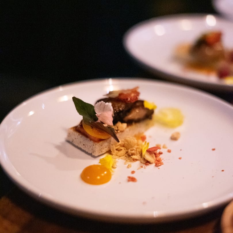 Gourmet appetizer on a white plate, featuring a small toast with toppings and edible flowers.