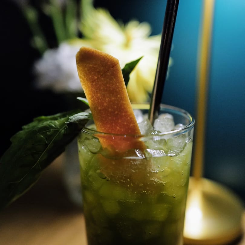 Green cocktail with ice, an orange peel garnish, and a dark straw, against a blue background.