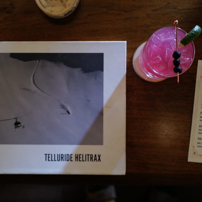 Pink cocktail with olives and lime on a wooden table next to a book titled "Telluride Helitrax".