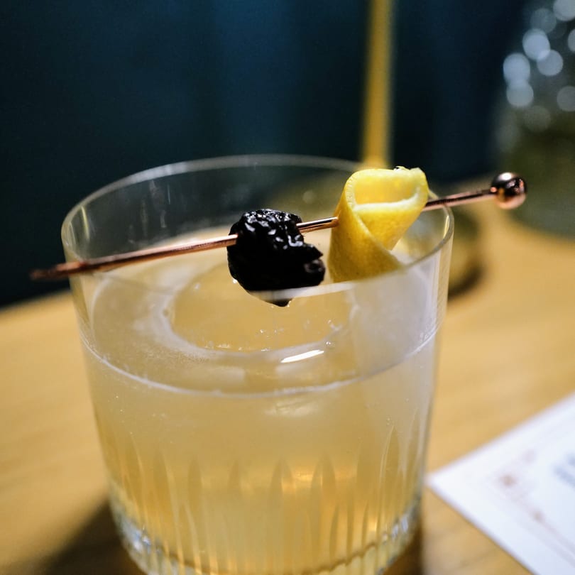 Close-up of a cocktail with a lemon twist and dark cherry on a copper skewer.