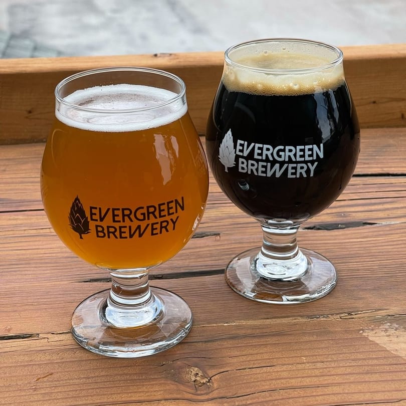 Two Evergreen Brewery glasses, one with light beer and one with dark beer, on a wooden table.