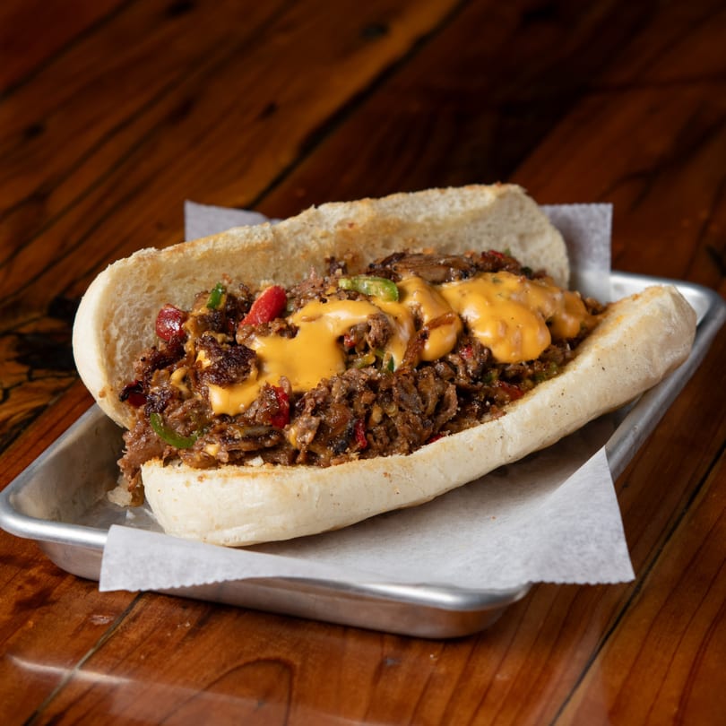 Cheesesteak sandwich with melted cheese, peppers, and onions on a metal tray at Evergreen Brewery.
