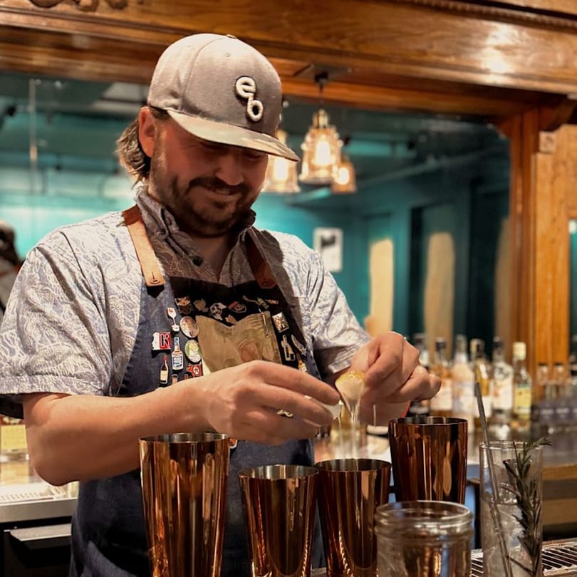 A smiling bartender in a hat and apron squeezes a citrus peel over copper shakers at a bar.