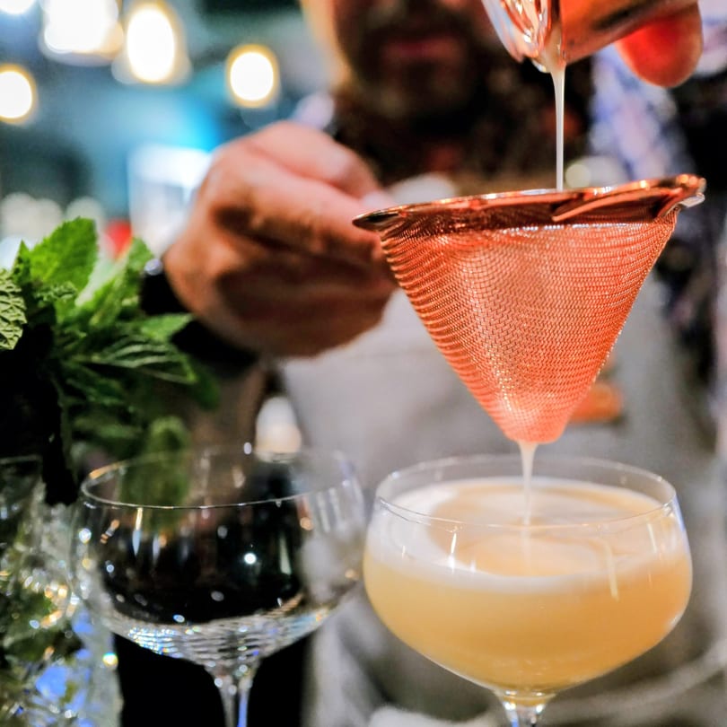 A bartender strains a creamy yellow cocktail into a coupe glass, with mint and another glass nearby.
