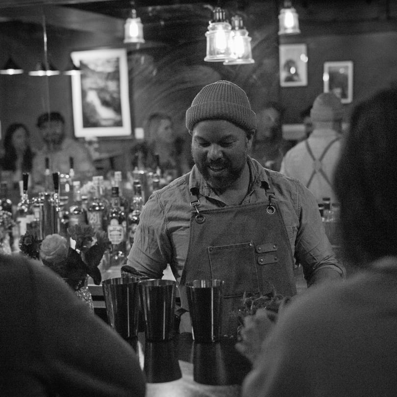 Smiling bartender in a beanie and apron, serving drinks at a lively bar with bottles in the background.