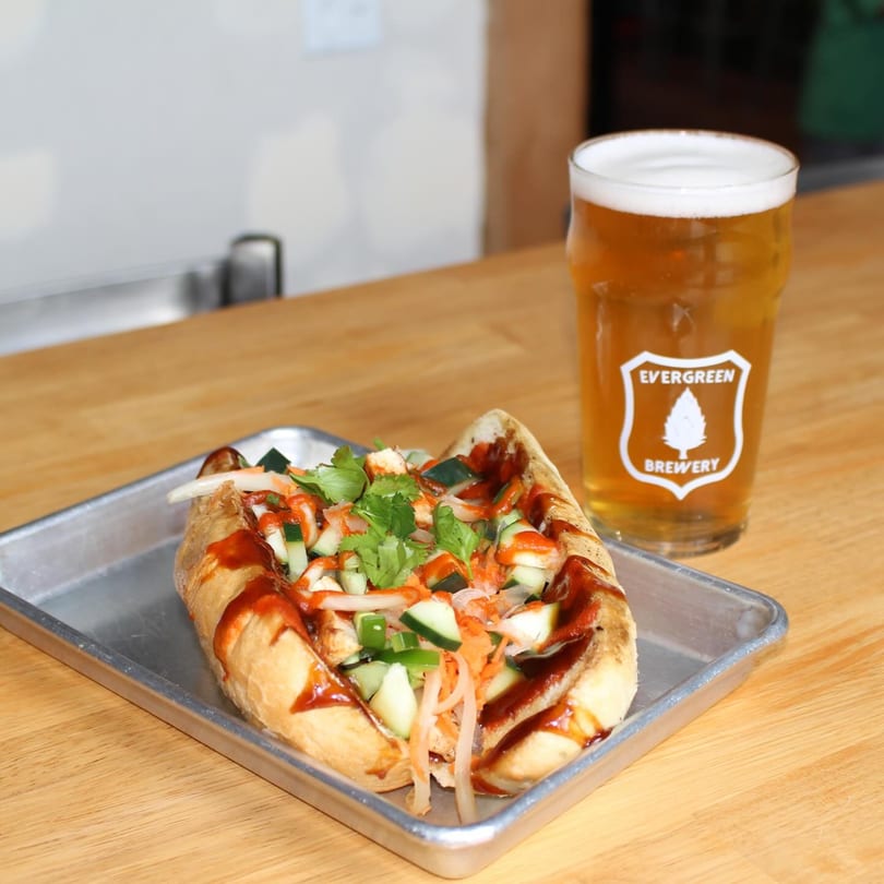 A banh mi sandwich with cilantro, cucumbers, and sauce, next to a pint of Evergreen Brewery beer.