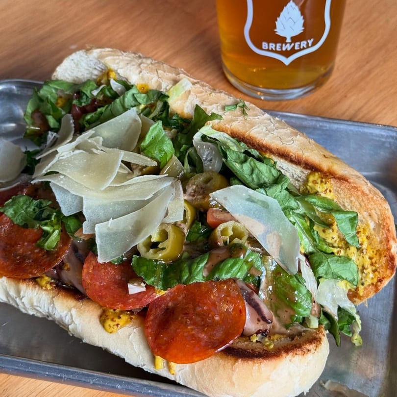A large sub sandwich with pepperoni, cheese, lettuce, and peppers, next to a glass of Evergreen Brewery beer.
