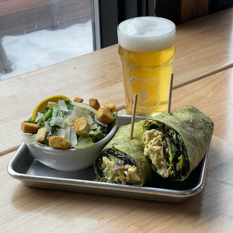 A chicken salad wrap, side salad, and a pint of Evergreen Brewery beer on a metal tray.