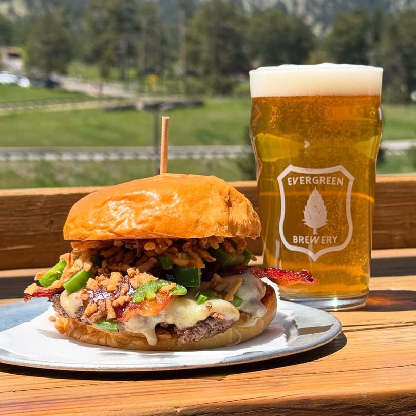 A burger with bacon, jalapeños, and crispy onions next to an Evergreen Brewery beer.