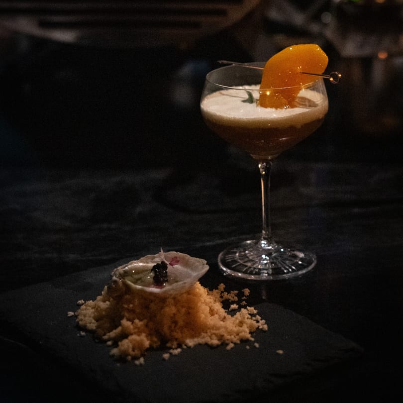A cocktail with orange garnish and a small dish of food on a dark surface.