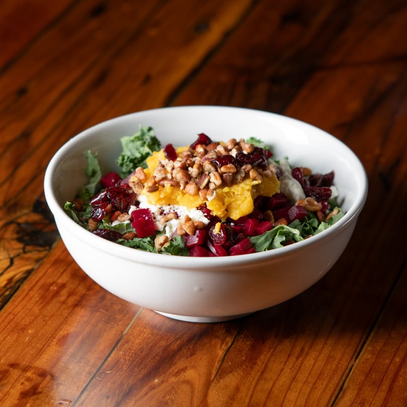 A vibrant salad with kale, beets, butternut squash, goat cheese, cranberries, and pecans in a white bowl.