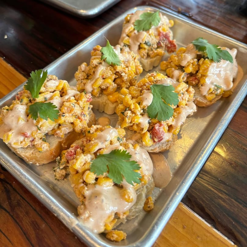 Six corn and tomato bruschetta bites with creamy sauce and cilantro on a metal tray.